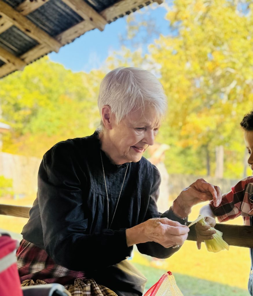 An elderly woman in a black period dress demonstrates crafting with a child at the Annual Harvest Festival in Heritage Village, Texas, surrounded by autumn foliage.
