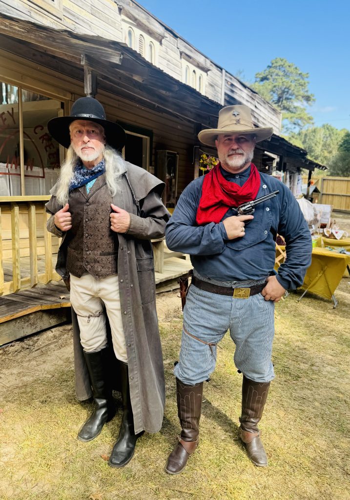 Two men in period costumes stand outdoors at a heritage festival, one wearing a black hat and coat, and the other a cowboy hat and red scarf, with a historic building in the background.