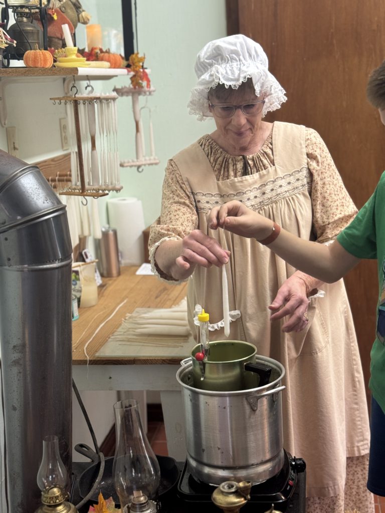 A woman in period dress teaches a child how to dip candles in a historic setting, surrounded by vintage decor and tools.