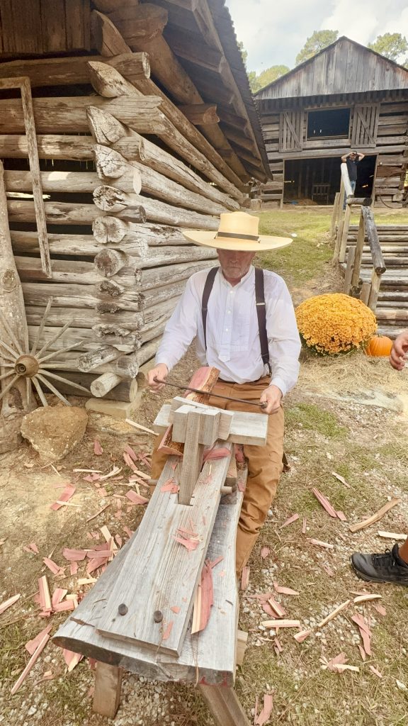 A craftsman in period attire works at a wooden bench, shaping wood in Heritage Village during the Annual Harvest Festival.